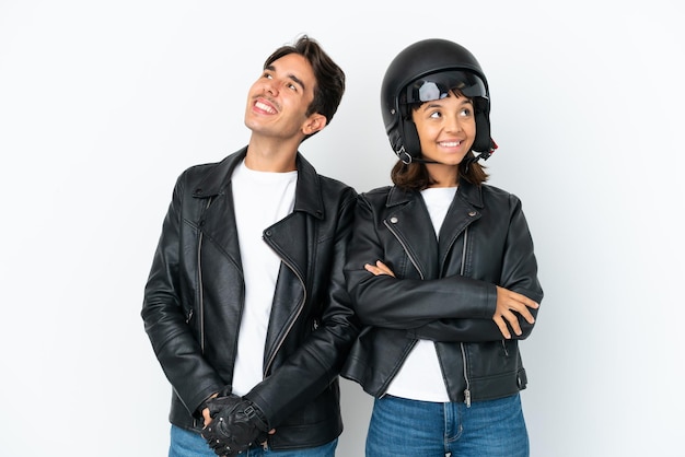 Jeune couple de race mixte avec un casque de moto isolé sur fond blanc levant les yeux en souriant
