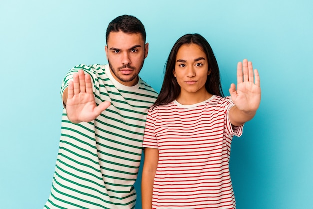 Jeune Couple De Race Mixte Isolé Sur Fond Bleu Debout Avec La Main Tendue Montrant Un Panneau D'arrêt, Vous Empêchant.