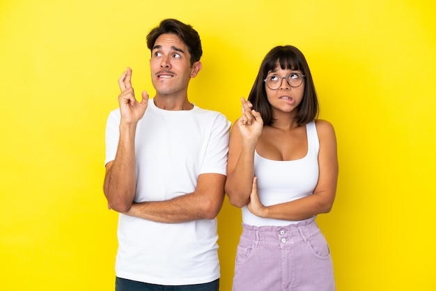 Jeune couple de race mixte isolé sur fond jaune avec les doigts croisés et souhaitant le meilleur