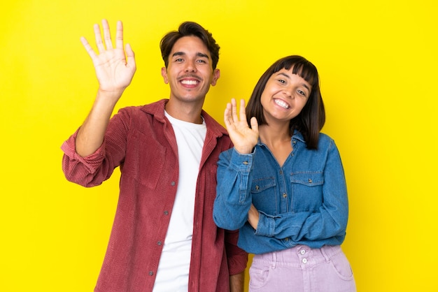 Jeune couple de race mixte isolé sur fond jaune saluant avec la main avec une expression heureuse