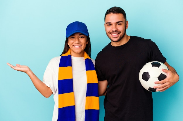 Jeune couple de race mixte nettoyant la maison isolée sur fond bleu montrant un espace de copie sur une paume et tenant une autre main sur la taille.