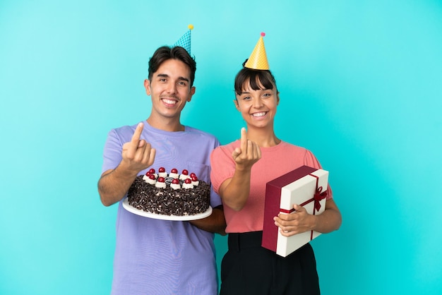 Jeune couple de race mixte tenant un gâteau d'anniversaire et un cadeau isolé sur fond bleu invitant à venir avec la main Heureux que vous soyez venu