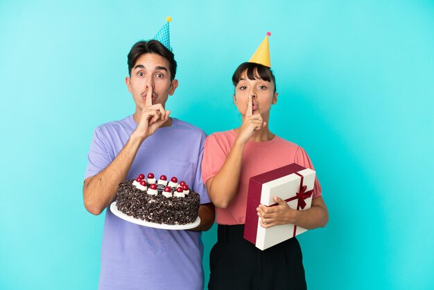 Jeune couple de race mixte tenant un gâteau d'anniversaire et un cadeau isolé sur fond bleu montrant un signe de geste de silence mettant le doigt dans la bouche