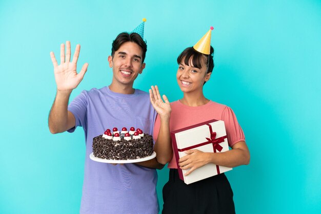 Jeune couple de race mixte tenant un gâteau d'anniversaire et un cadeau isolé sur fond bleu saluant avec la main avec une expression heureuse