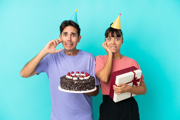 Jeune couple de race mixte tenant un gâteau d'anniversaire et présent isolé sur fond bleu couvrant les deux oreilles avec les mains