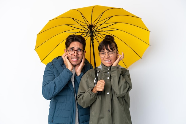 Jeune couple de race mixte tenant un parapluie isolé sur fond blanc couvrant les deux oreilles avec les mains