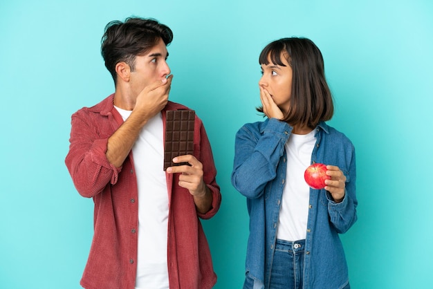 Jeune couple de race mixte tenant une pomme et du chocolat isolé sur fond bleu couvrant la bouche avec les mains pour avoir dit quelque chose d'inapproprié