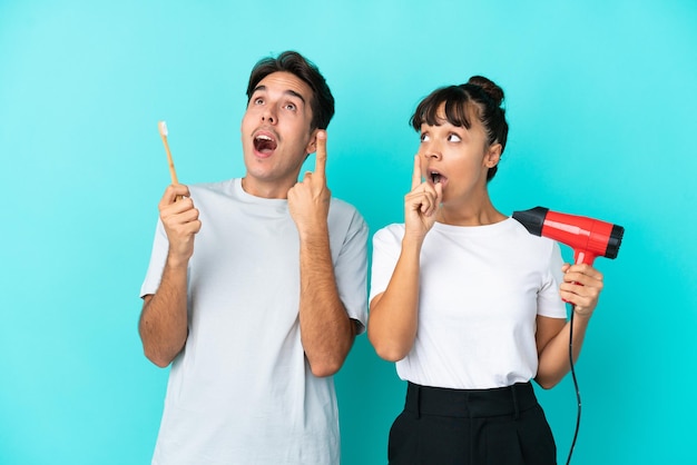 Jeune couple de race mixte tenant un sèche-cheveux et une brosse à dents isolé sur fond bleu pensant une idée pointant le doigt vers le haut