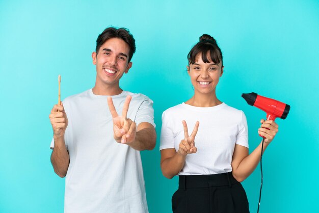 Jeune couple de race mixte tenant un sèche-cheveux et une brosse à dents isolé sur fond bleu souriant et montrant le signe de la victoire