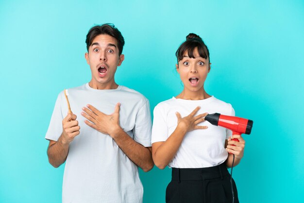 Jeune couple de race mixte tenant un sèche-cheveux et une brosse à dents isolé sur fond bleu surpris et choqué en regardant à droite