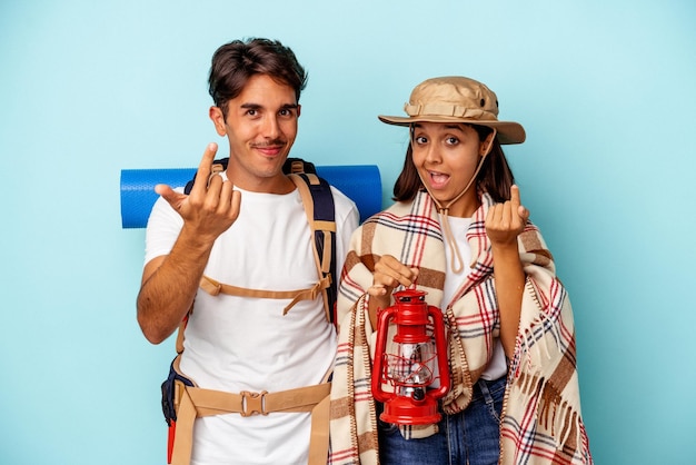 Jeune couple de randonneurs de race mixte isolé sur fond bleu pointant du doigt vers vous comme s'il vous invitait à vous rapprocher.