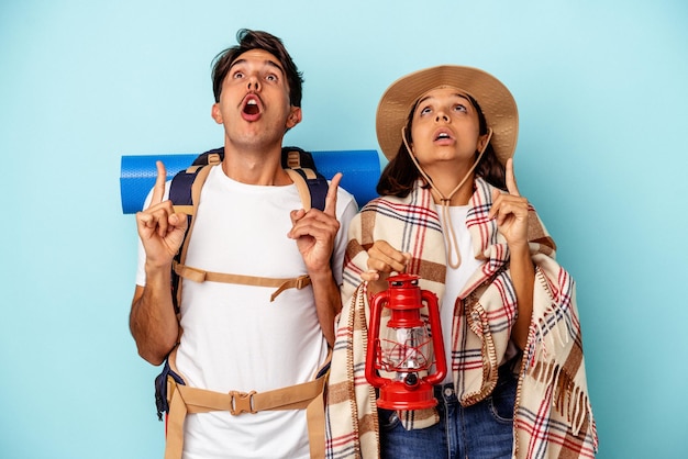 Jeune couple de randonneurs de race mixte isolé sur fond bleu pointant vers le haut avec la bouche ouverte.