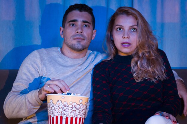 Jeune couple regardant un film thriller dans une pièce sombre avec du pop-corn sur le canapé pleinement attention