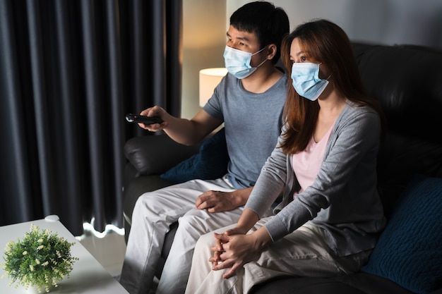 Jeune couple regardant la télévision sur un canapé et portant un masque médical pour protéger le coronavirus (Covid-19)