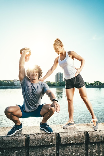 Jeune couple de remise en forme fait de l'exercice avec kettlebell sur le mur au bord de la rivière au coucher du soleil. L'homme est accroupi et tient un kettlebell, et la femme le soutient.