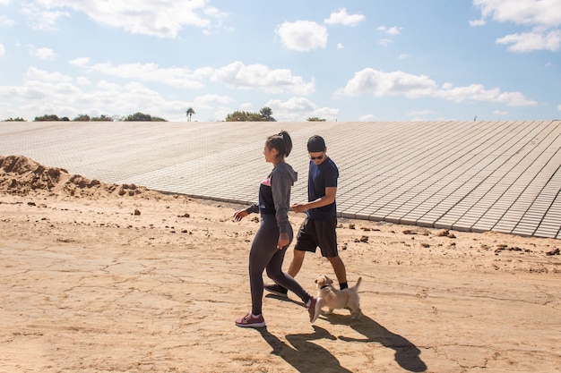 Jeune couple de remise en forme marchant dans le sable pendant que leur chiot les suit.