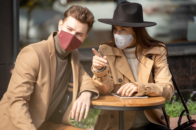 Jeune couple reposant dans des masques de protection et des vêtements décontractés intelligents assis près d'une petite table ronde et regardant quelque chose que cette fille pointe du doigt