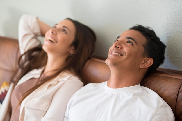 Jeune couple rêvant ensemble à la maison assis sur le canapé et levant les yeux avec un sourire Personnes vivant dans une nouvelle maison après avoir déménagé et prêt bancaire hypothécaire Concept de garçons et vie future dans un appartement