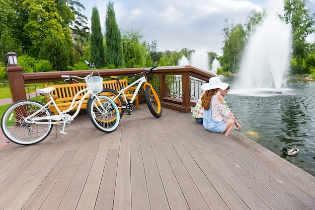 Jeune couple romantique assis sur la terrasse en bois après avoir fait du vélo et nourri les canards dans un étang dans un parc avec fontaine et différents arbres en arrière-plan