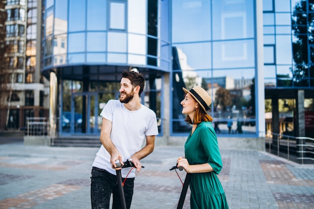 Un jeune couple romantique avec des scooters électriques à une date, marchant dans la ville.