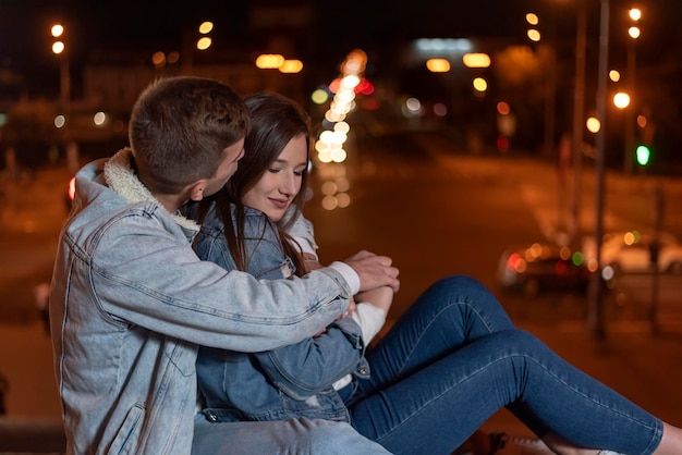 Le jeune couple s'assied embrassant sur le fond de ville de soirée