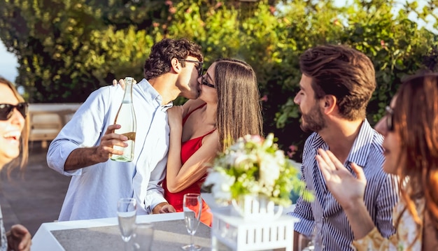 Jeune couple s'embrassant au restaurant fête de célébration d'un engagement avec des amis des gens qui s'amusent à boire du vin blanc