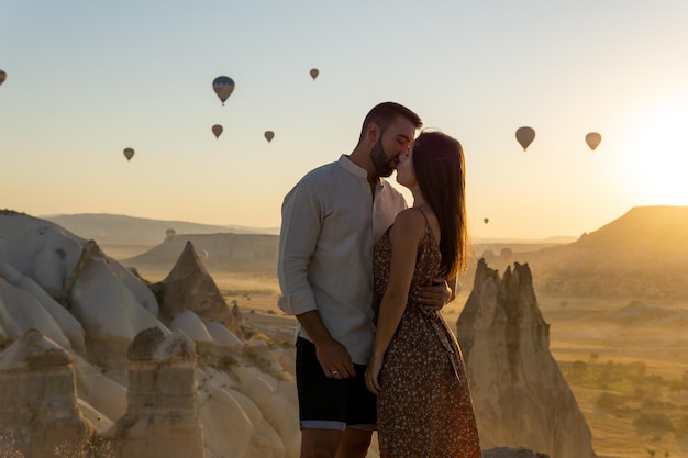 Jeune couple s'embrassant très amoureux au premier plan en regardant les montgolfières typiques de la région de la cappadoce volant au lever du soleil