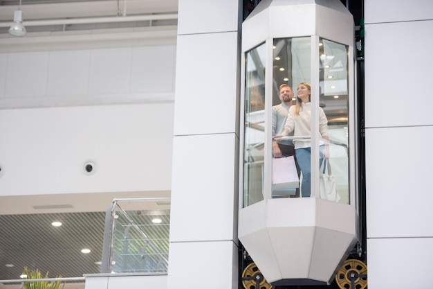 Jeune couple avec des sacs en papier debout dans l'ascenseur tout en se déplaçant vers le haut à l'intérieur d'un grand centre commercial contemporain pendant le shopping