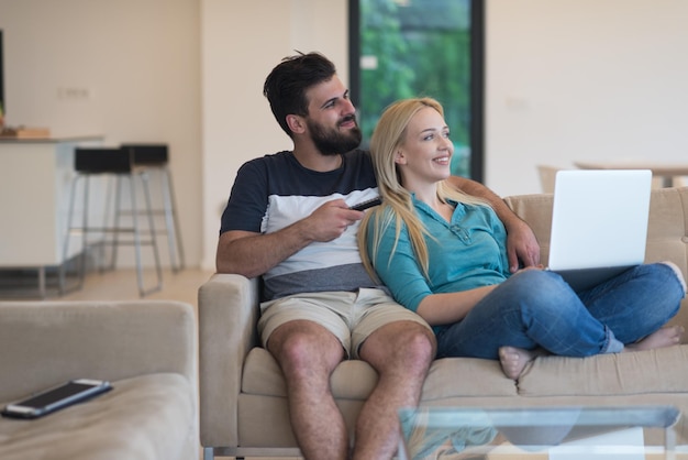 Un jeune couple se détend sur le canapé du salon de luxe, à l'aide d'un ordinateur portable et d'une télécommande