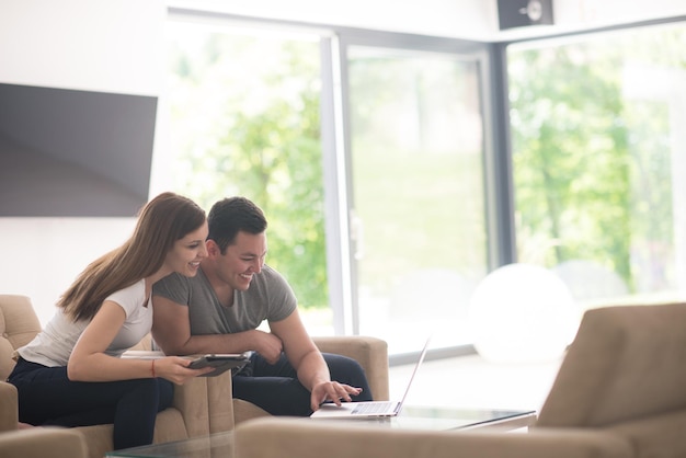 Jeune couple se relaxant dans une maison luxueuse avec des ordinateurs tablettes et portables lisant dans le salon sur le canapé-lit.