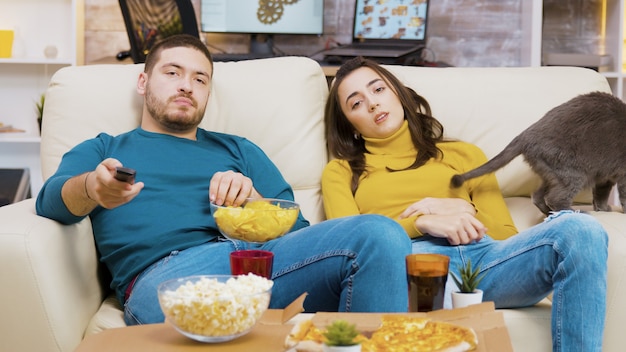 Jeune couple se reposant sur le canapé avec son chat devant la télévision. Homme mangeant des chips.