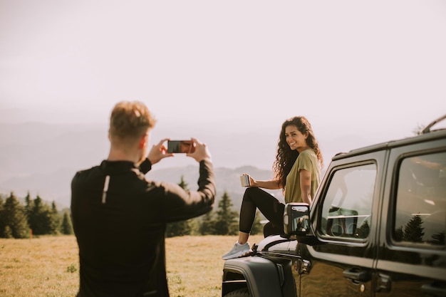 Jeune couple se reposant sur un capot de véhicule tout-terrain et prenant des photos avec un téléphone portable à la campagne