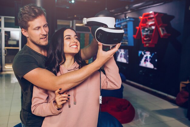 Jeune couple se tenir ensemble dans la salle de jeux et tenir des lunettes VR. Ils le regardent. Sourire de jeune femme.