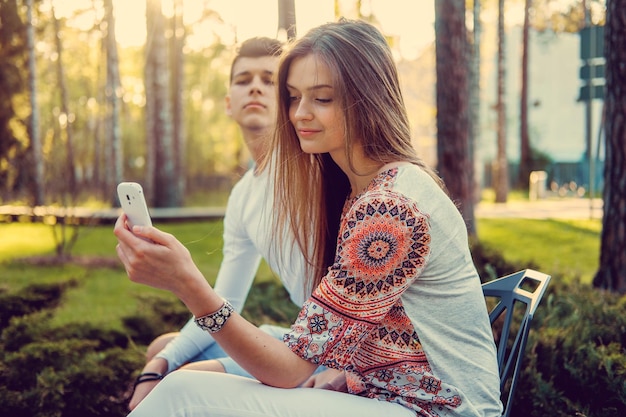 Jeune couple séduisant assis sur un banc dans un parc et utilisant un smartphone.