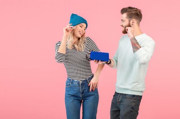 Jeune couple séduisant écoutant de la musique sur un haut-parleur sans fil portant une tenue élégante et cool souriante de bonne humeur positive posant sur fond rose