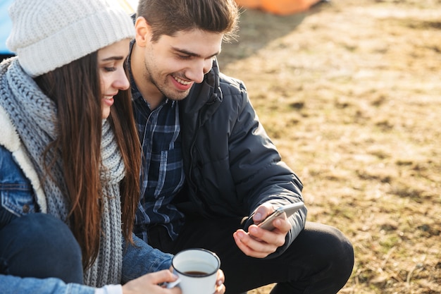 Jeune couple séduisant se reposant assis à la tente à l'extérieur, tenant une tasse et un thermos, utilisant un téléphone portable