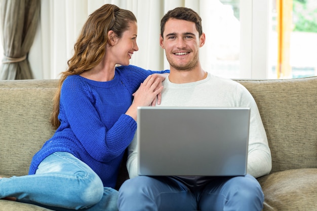Jeune couple souriant face à face sur le canapé et utilisant un ordinateur portable dans le salon