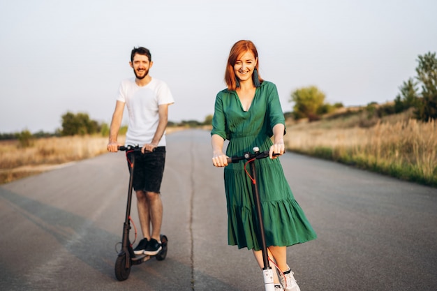 Jeune couple souriant heureux s'amuser au volant de scooter électrique sur la route à la campagne. Technologies de contenu