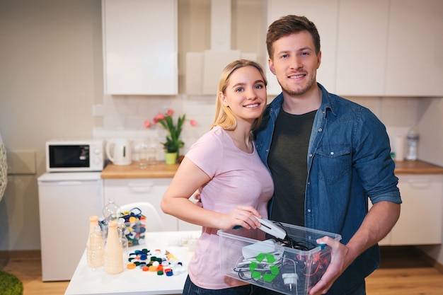 Jeune couple souriant tenant un conteneur de recyclage rempli de déchets électroniques