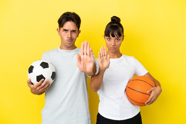 Jeune couple sportif jouant au football et au basket-ball isolé sur fond jaune faisant un geste d'arrêt niant une situation qui pense mal