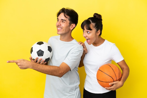 Jeune couple sportif jouant au football et au basket-ball isolé sur fond jaune pointant vers le côté pour présenter un produit