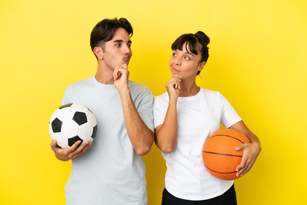 Jeune couple sportif jouant au football et au basket-ball isolé sur fond jaune regardant les uns les autres