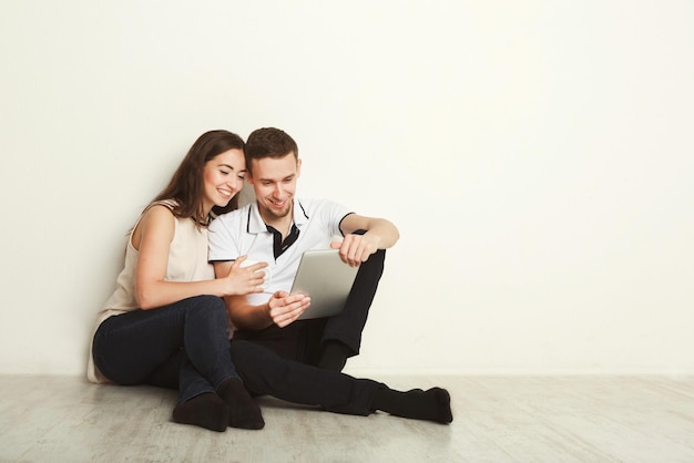 Jeune couple surfant sur le web sur tablette numérique assis sur le sol dans le salon vide du nouvel appartement sur fond blanc, espace pour copie