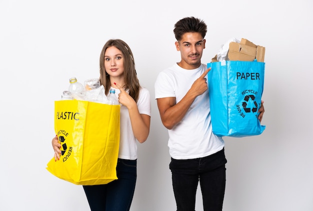 Jeune couple tenant un sac plein de plastique et de papier sur blanc pointant vers le côté pour présenter un produit
