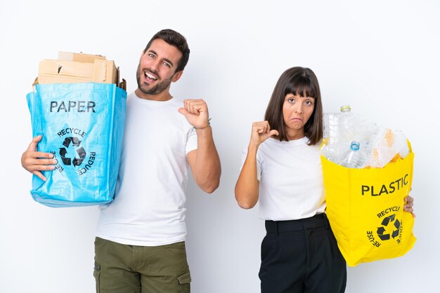 Jeune couple tenant un sac plein de plastique et de papier à recycler isolé sur fond blanc fier et satisfait de vous-même concept d'amour