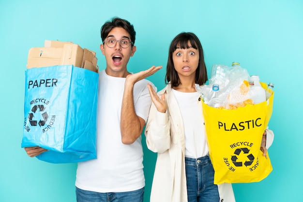 Jeune couple tenant un sac plein de plastique et de papier à recycler isolé sur fond bleu avec une expression faciale surprise et choquée