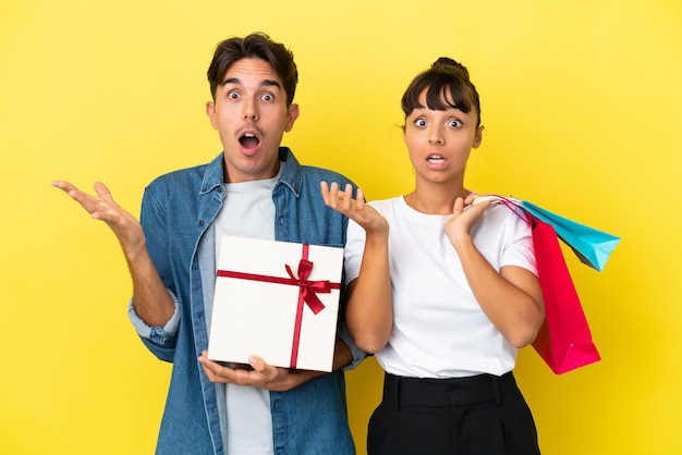 Jeune couple tenant des sacs à provisions et présent isolé sur fond jaune avec une expression faciale surprise et choquée