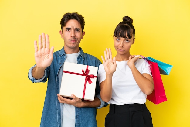 Jeune couple tenant des sacs à provisions et présent isolé sur fond jaune faisant un geste d'arrêt niant une situation qui pense mal