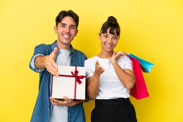 Jeune couple tenant des sacs à provisions et présent isolé sur fond jaune se serrant la main pour conclure une bonne affaire