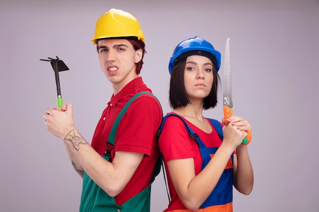 Jeune couple en uniforme de travailleur de la construction et casque de sécurité debout dos à dos mec agressif tenant une fille sérieuse tenant une houe tenant la main a vu les deux regardant la caméra isolée sur un mur blanc